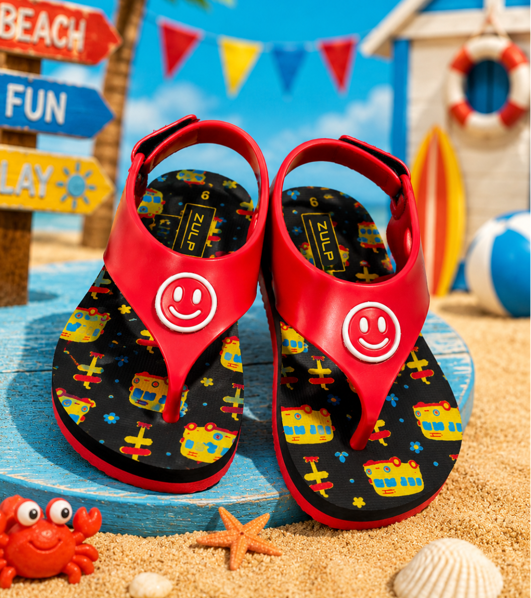 A pair of red flip-flop sandals with black patterned soles featuring colorful airplanes and a white smiley face logo on the toe strap, displayed on a blue wooden platform in a beach-themed setting with sand, a crab toy, and a starfish.